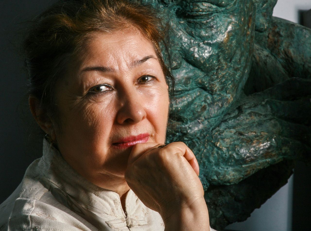 Colour portrait of Munira Shahidi, her chin resting on her hand, beside a sculpted bronze head with a green patina.
